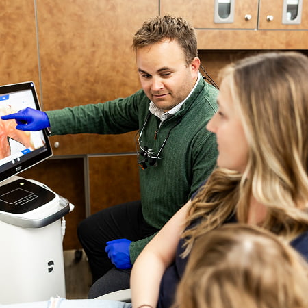 Dr. Kyle Burgmeier smiling while explaining a dental procedure to a female patient and her son using a monitor at Burgmeier Dentistry