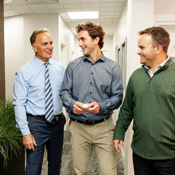 Dr. Dan Burgmeier, Dr. Kyle Burgmeier, and Dr. Joe Burgmeier having a friendly discussion while holding a denture model in a clean dental hallway at Burgmeier Dentistry